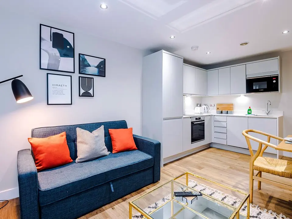 Living area with a sofa and orange cushions, a glass coffee table and a modern white kitchen behind